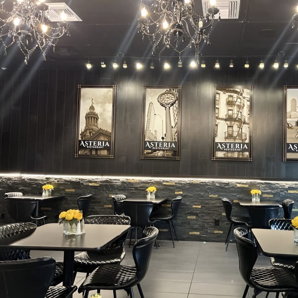 Interior of Asteria Restaurant with black tables, chairs, chandeliers, and framed city prints on dark walls.