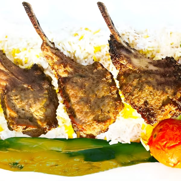 Three grilled lamb chops with rice, a roasted tomato, and green garnish on a white plate.